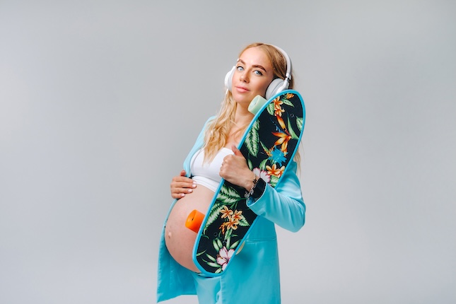 pregnant-girl-turquoise-suit-with-skateboard-her-hands-headphones-stands-gray-background